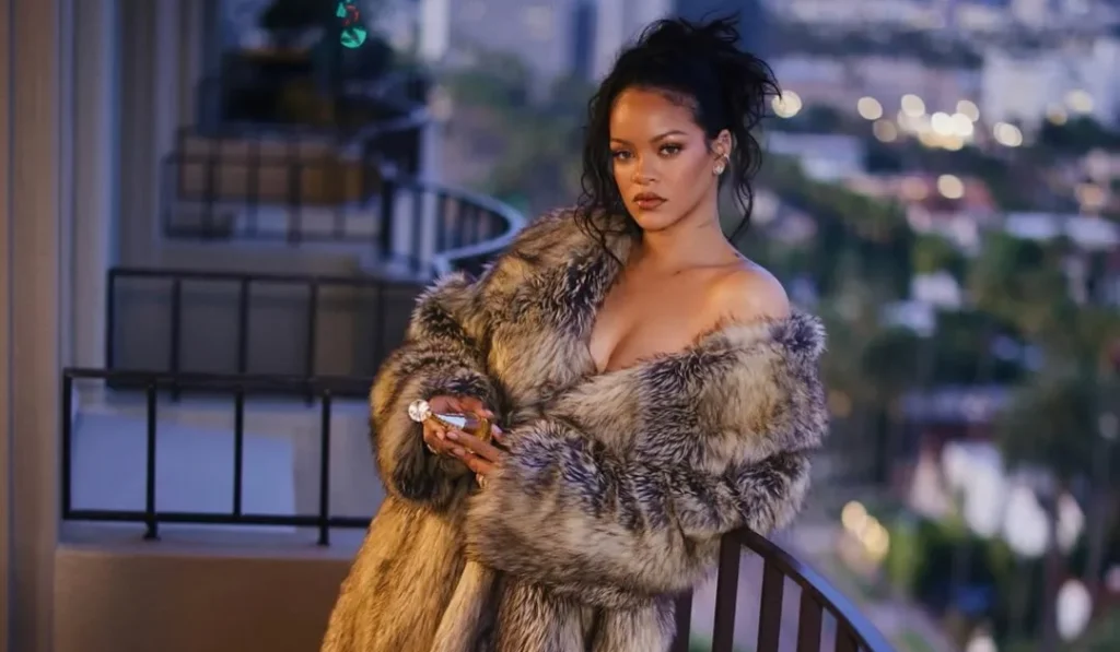 Rihanna on a balcony at night in a glamorous, long, off-the-shoulder faux fur coat and high heels, overlooking a city.