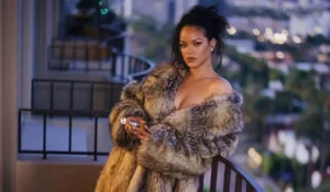 Rihanna on a balcony at night in a glamorous, long, off-the-shoulder faux fur coat and high heels, overlooking a city.