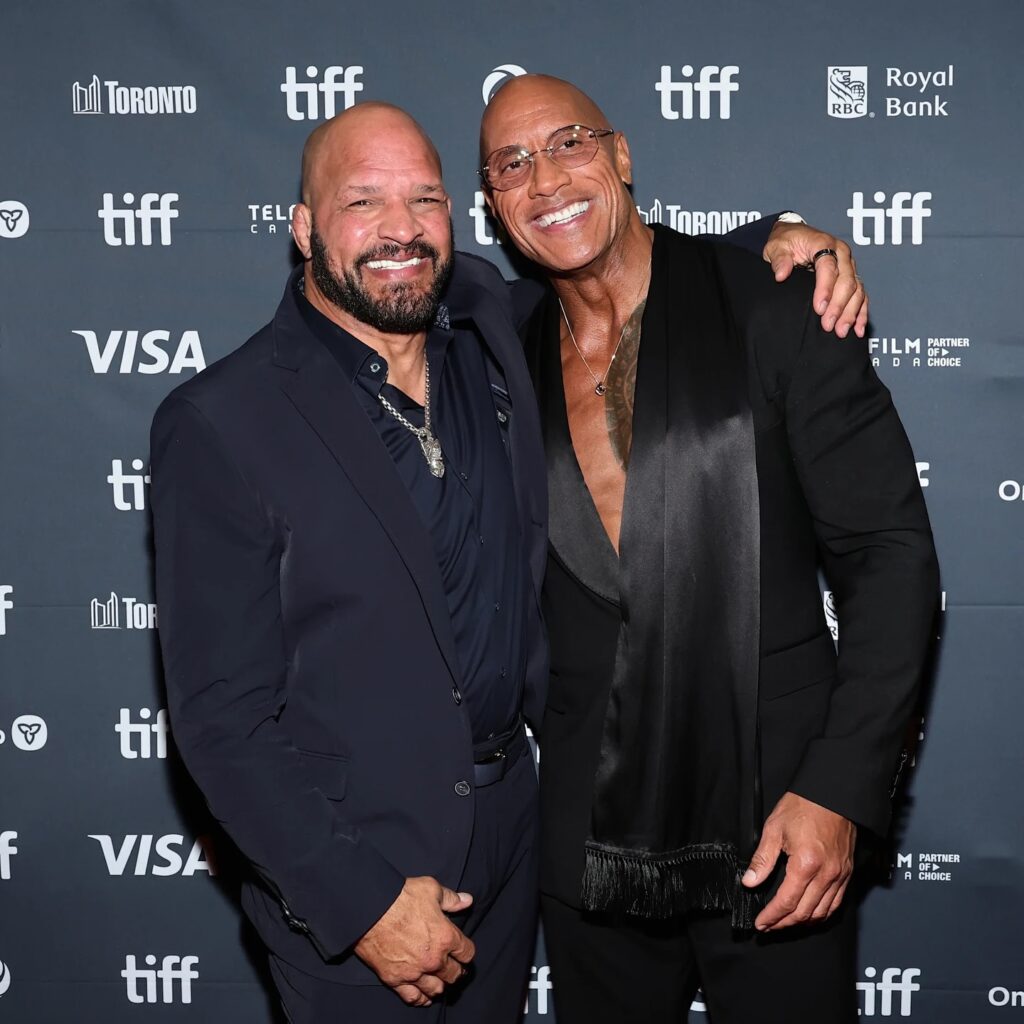 Mark Kerr and Dwyane Johnson in formal attire pose together on a TIFF step-and-repeat backdrop, one wearing glasses and scarf-like lapel detail.