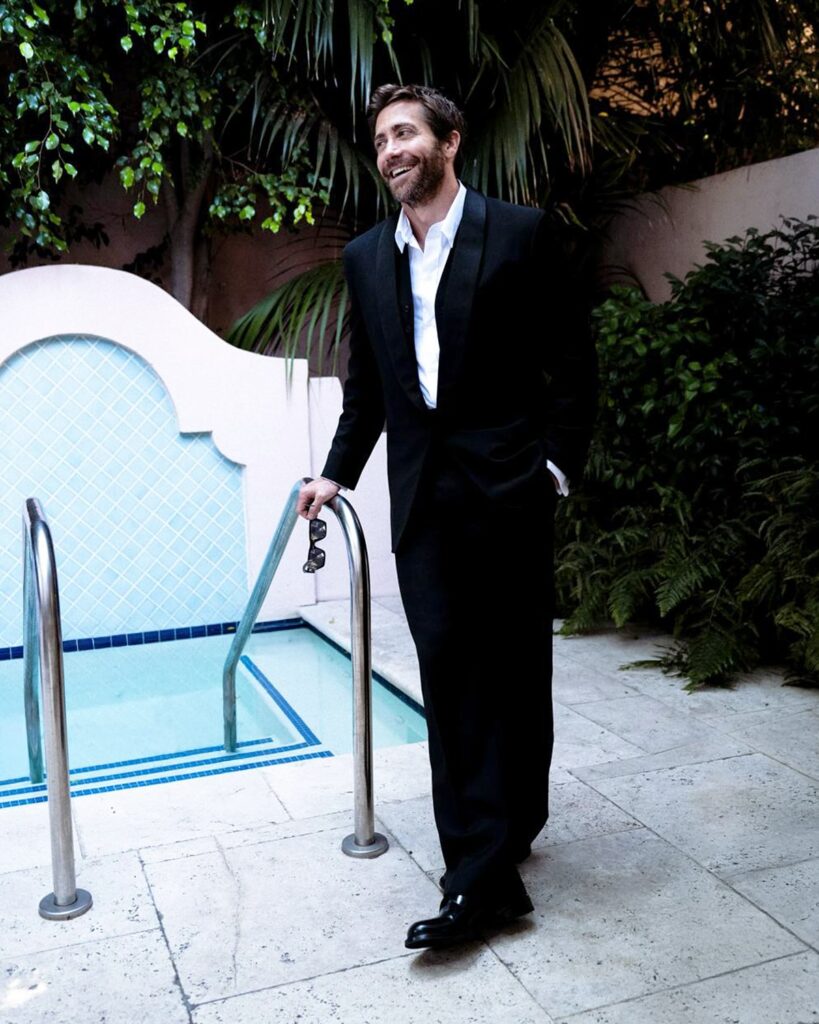 Jake Gyllenhaal in a black tuxedo stands by a small tiled pool, smiling with a relaxed posture. He holds sunglasses, one hand in pocket, surrounded by lush greenery and stucco walls, creating an elegant, candid outdoor setting at dusk.