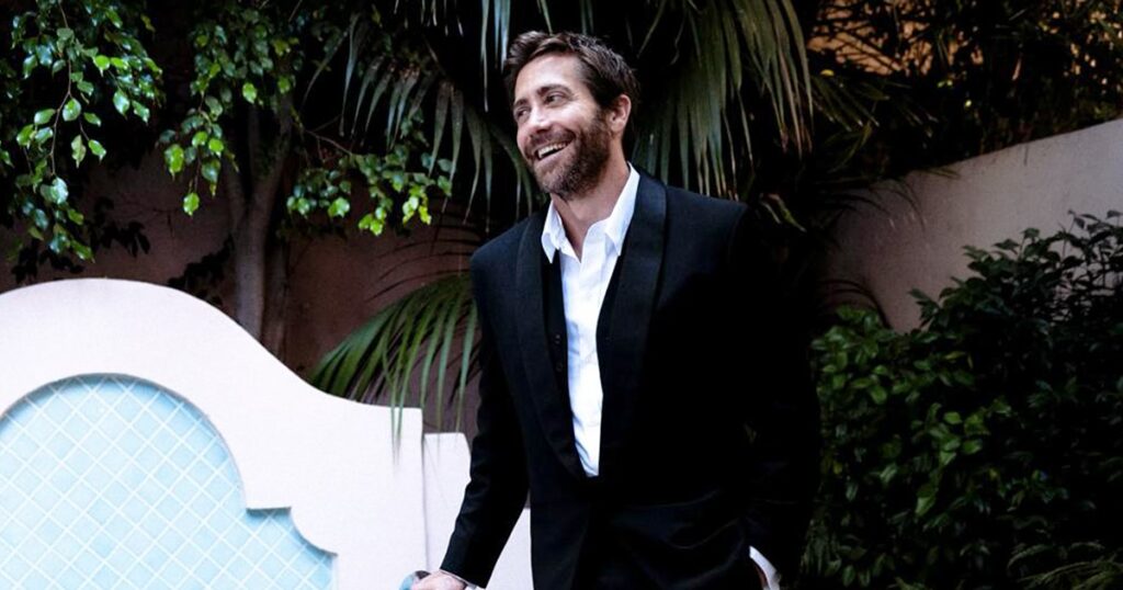 Jake Gyllenhaal in a black tuxedo stands by a small tiled pool, smiling with a relaxed posture. He holds sunglasses, one hand in pocket, surrounded by lush greenery and stucco walls, creating an elegant, candid outdoor setting at dusk.