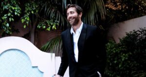 Jake Gyllenhaal in a black tuxedo stands by a small tiled pool, smiling with a relaxed posture. He holds sunglasses, one hand in pocket, surrounded by lush greenery and stucco walls, creating an elegant, candid outdoor setting at dusk.