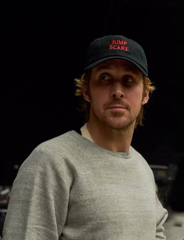 Ryan Gosling wearing a black cap with red "JUMP SCARE" text and a grey sweatshirt, looking to the side.