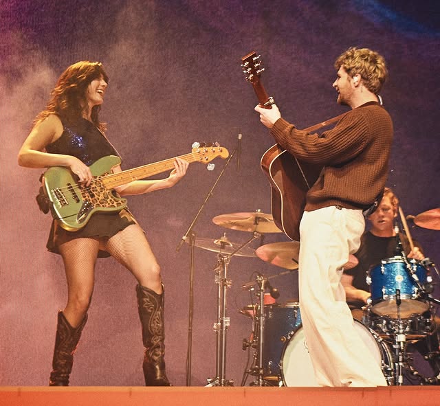 Three musicians on stage: a smiling bassist in boots playing a green bass, a guitarist facing them with an acoustic guitar, and a drummer behind a kit under warm stage lights.