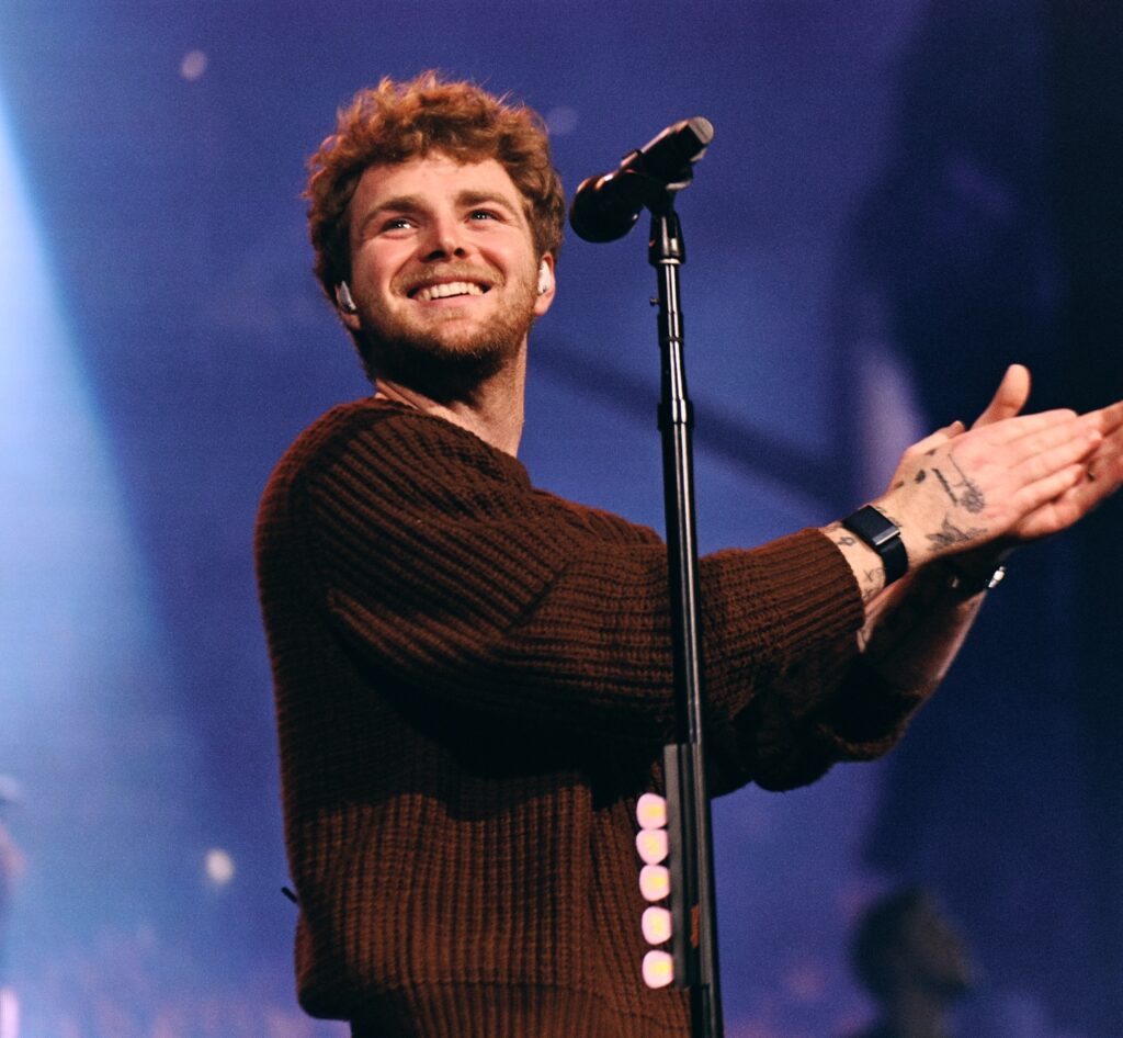 Alex Warren at a microphone smiling and clapping on stage, wearing a brown sweater, with warm spotlights and a blurred audience behind.