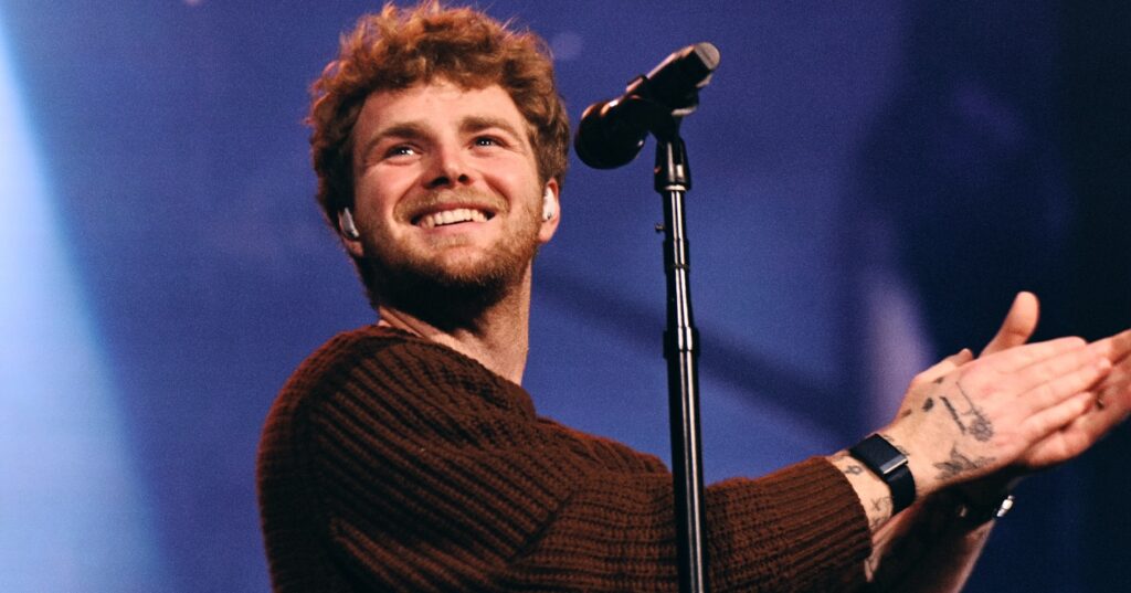 Singer Alex Warren at a microphone smiling and clapping on stage, wearing a brown sweater, with warm spotlights and a blurred audience behind.