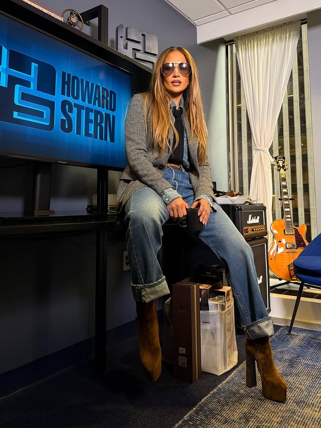 Woman seated in a studio setting wearing sunglasses, cuffed jeans, blazer, and platform boots beside a screen and guitar.