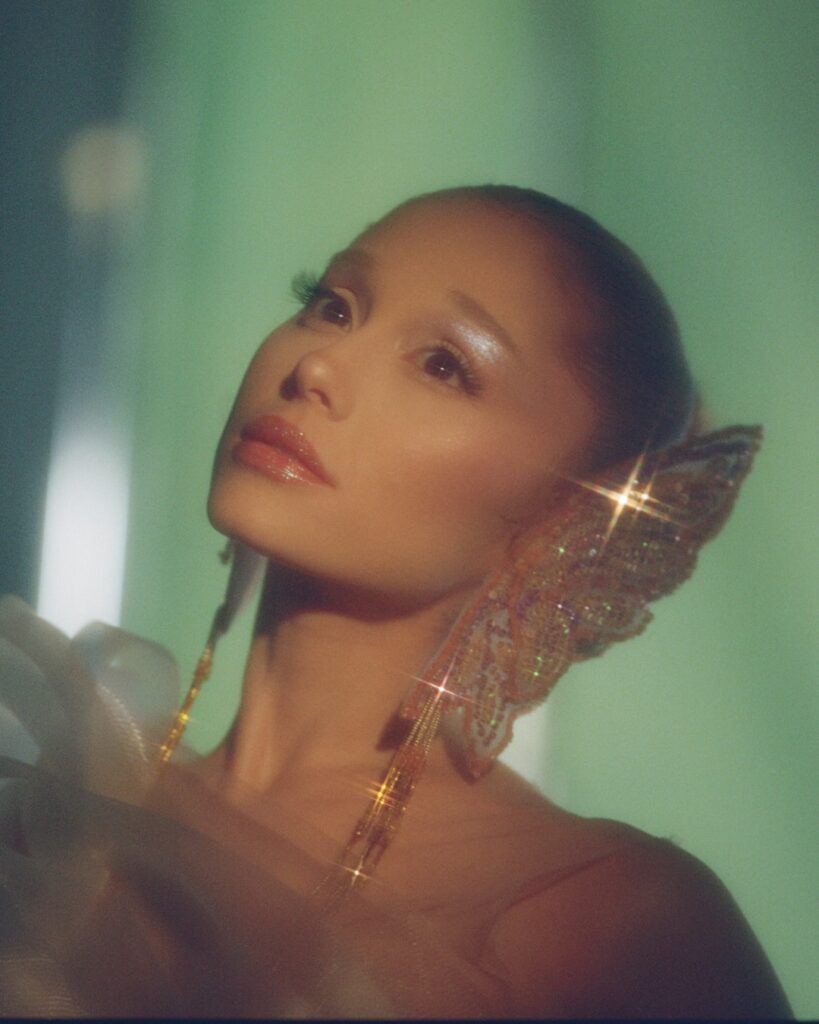 Close-up portrait of a woman with glossy makeup and shimmering wing-shaped beaded ear adornment, gazing upward in soft, dreamy lighting against a green background.
