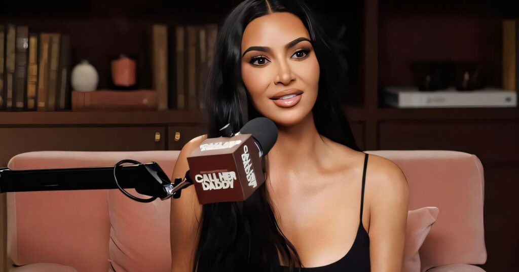 Kim Kardashian sits on a blush pink chair during a podcast interview, speaking into a “Call Her Daddy” microphone with a confident smile, surrounded by bookshelves in a cozy, warmly lit studio.