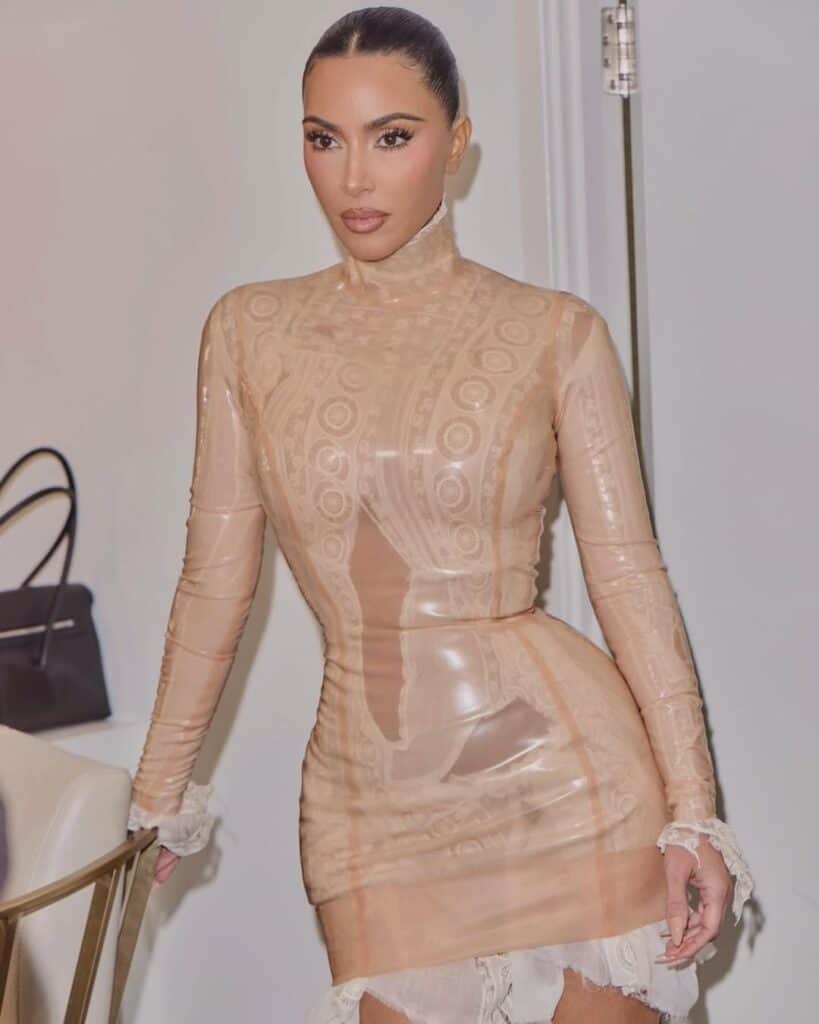 Kim Kardashian poses in a form-fitting beige latex dress with intricate lace patterns, showcasing her signature sleek style.