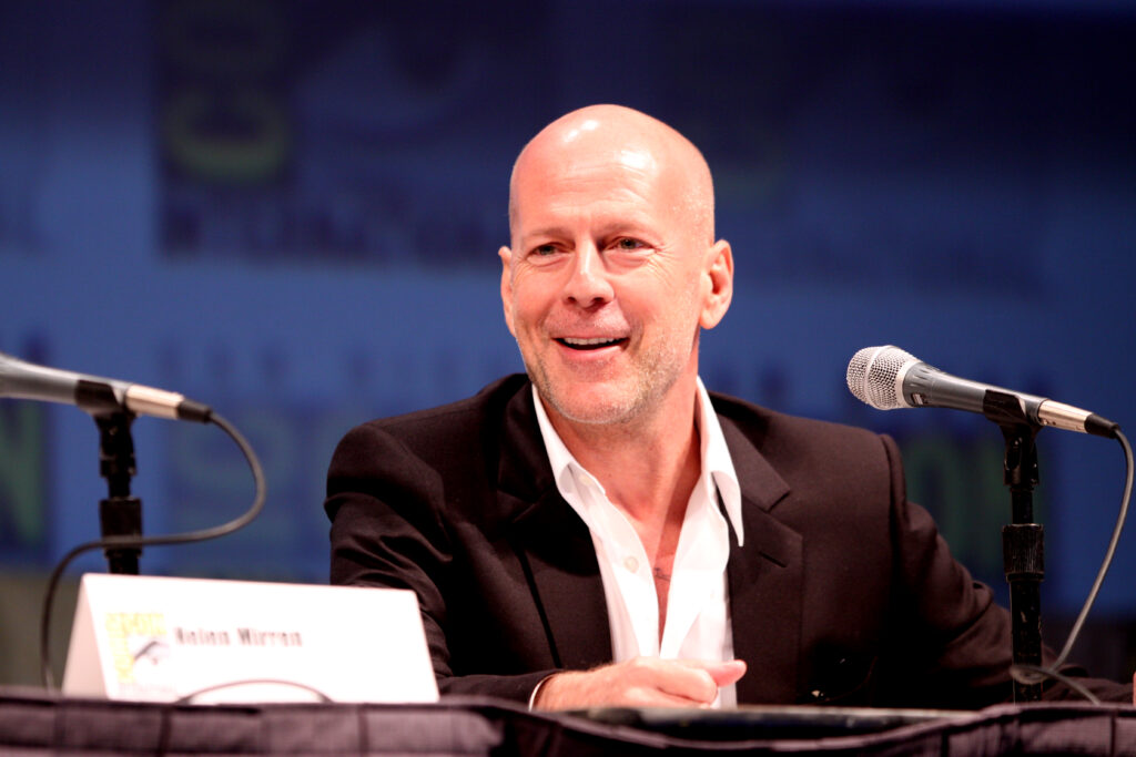 Bruce wills Smiling in a black suit and open-collar white shirt sits at a panel table between two microphones, speaking to an audience under warm stage lights with a blurred convention backdrop.