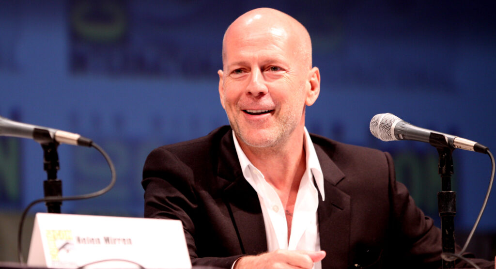 Bruce Wills in a black suit and open-collar white shirt sits at a panel table between two microphones, speaking to an audience under warm stage lights with a blurred convention backdrop.