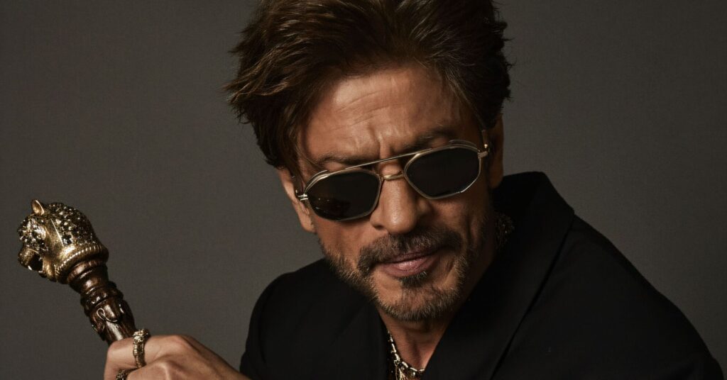 Shah Rukh Khan in a black suit, sunglasses, and a watch, holding a lion-head cane while wearing multiple gold rings and a heavy gold necklace.