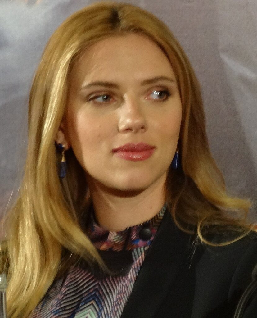 scarlett johansson with long blonde hair at a press conference, wearing patterned top and earrings.