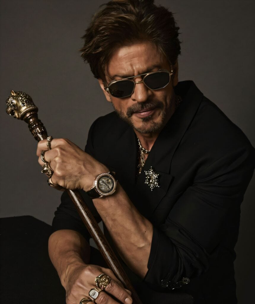 Shah Rukh Khan poses in a black suit and sunglasses, holding a gold lion-headed cane. He wears multiple statement rings, a luxury watch, and a jeweled brooch, exuding a powerful, stylish aura against a dark grey background.