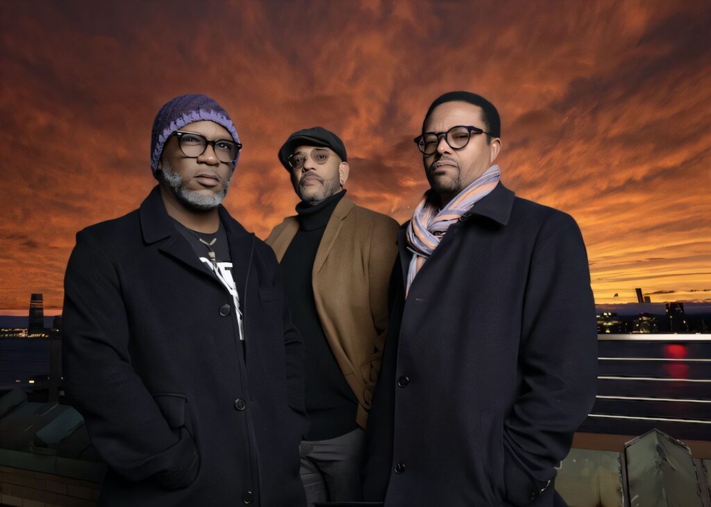Three middle-aged men stand on a waterfront at sunset, wearing winter coats, hats, and scarves, facing the camera with serious expressions against a dramatic orange sky.