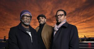 Three middle-aged men stand on a waterfront at sunset, wearing winter coats,