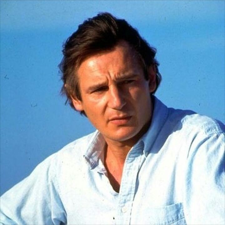 Liam Neeson in a black turtleneck and coat, smiling slightly, with a toothpick in his mouth at an event.