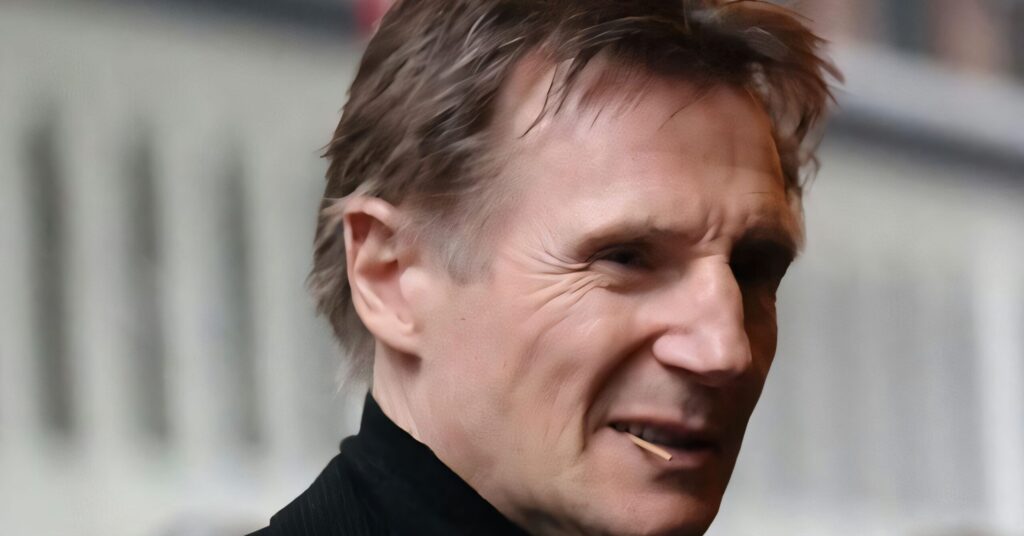 Headshot of a younger Liam Neeson outdoors, wearing a light blue button-down shirt against a clear blue sky.