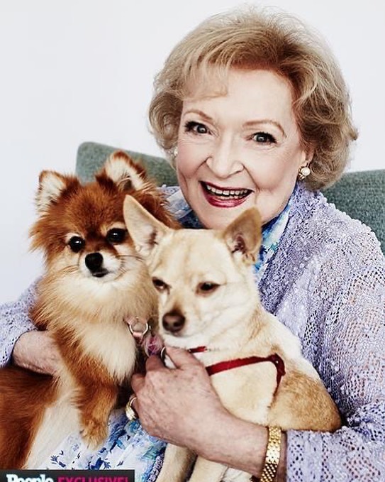 A color photo of a young Betty White sitting on porch steps with a fluffy tan Pekingese dog in her lap and a black dog at her feet.