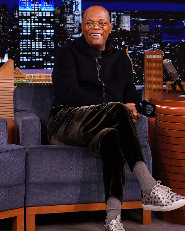 Smiling bald man in glasses seated on a late-night show couch, wearing a dark zip-up top and patterned sneakers.