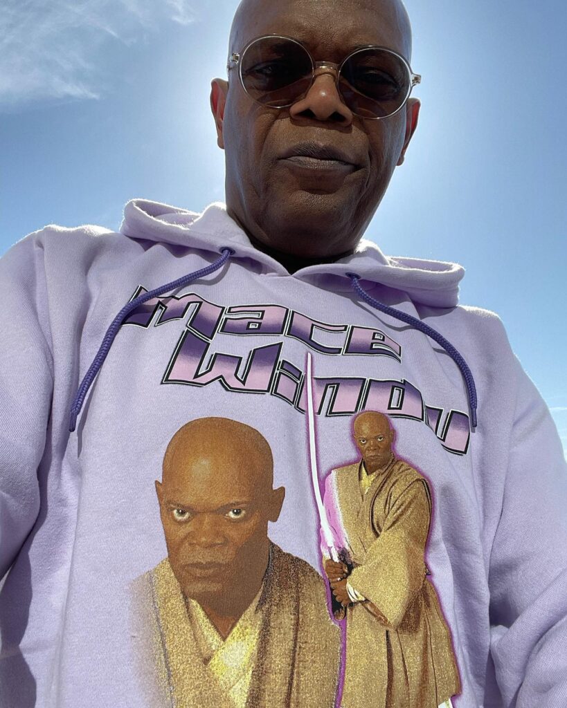  Selfie of a bald man wearing sunglasses and a lavender hoodie printed with vintage sci-fi character images, sky behind him.