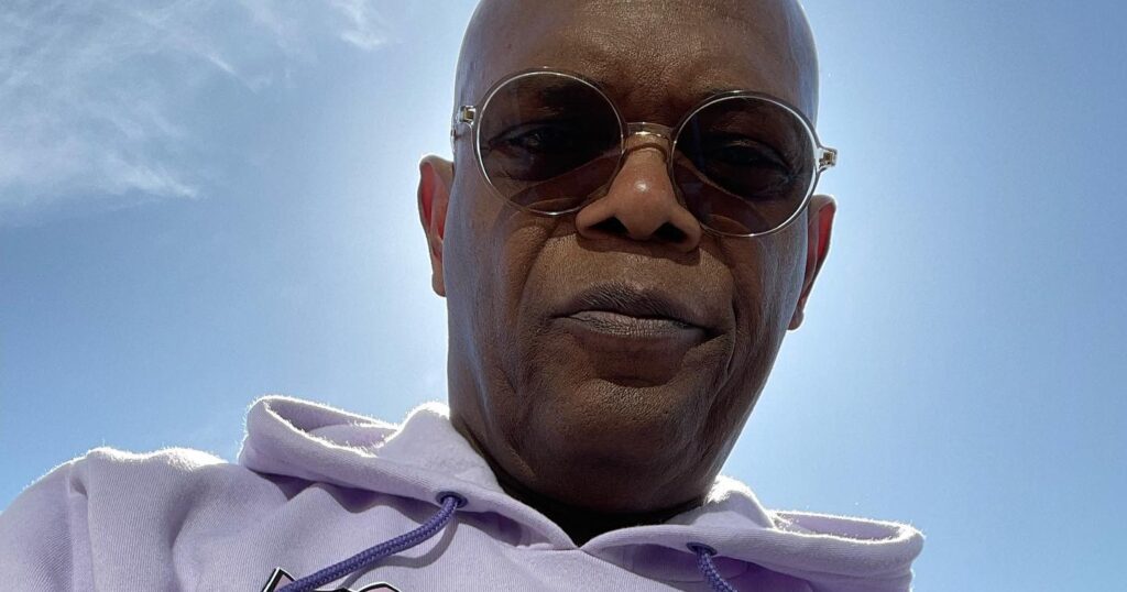 Selfie of Samuel L. Jackson wearing sunglasses and a lavender hoodie printed with vintage sci-fi character images, sky behind him.