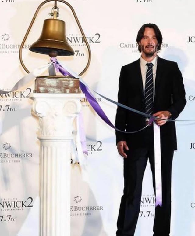Reeves stands next to a large bell on a white pillar, wearing a black suit and striped tie, pulling a white ribbon at a red carpet event.