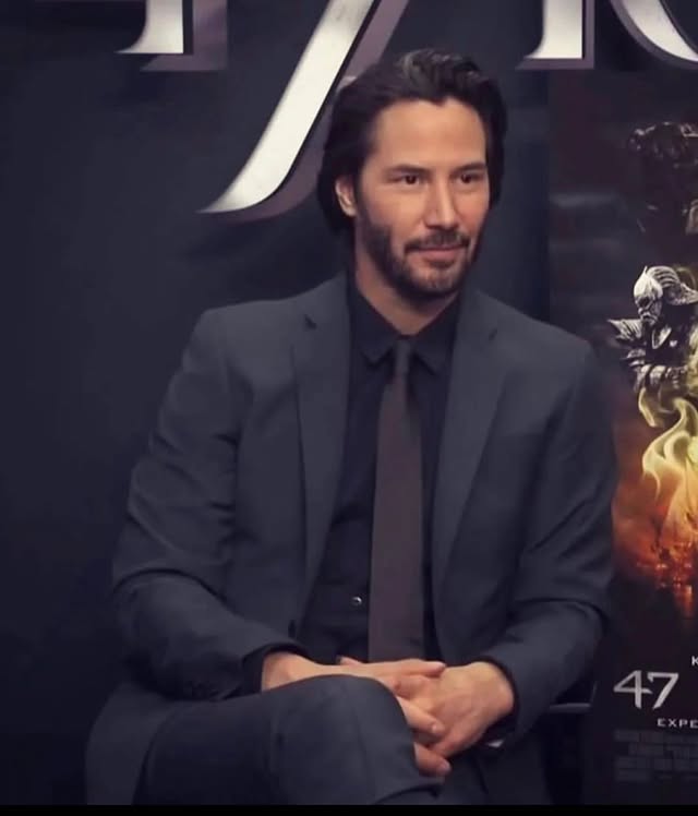 A portrait of actor Keanu Reeves seated, wearing a dark gray suit, black shirt, and tie, smiling subtly against a dark promotional backdrop.