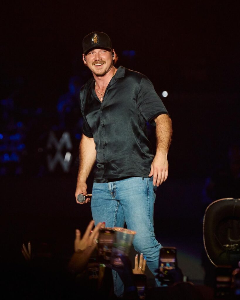 Country music star Morgan Wallen smiles on stage while wearing a black satin short-sleeved button-up shirt, blue jeans, and a dark baseball cap.