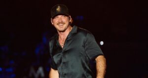 Country music star Morgan Wallen smiles on stage while wearing a black satin short-sleeved button-up shirt, blue jeans, and a dark baseball cap.