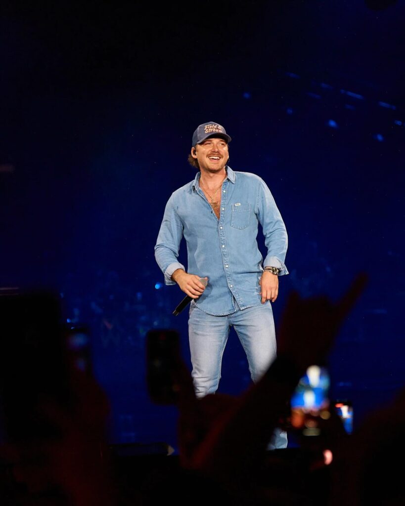 Country music singer Morgan Wallen performs on stage, smiling while dressed in a light-wash denim button-up shirt and matching jeans.