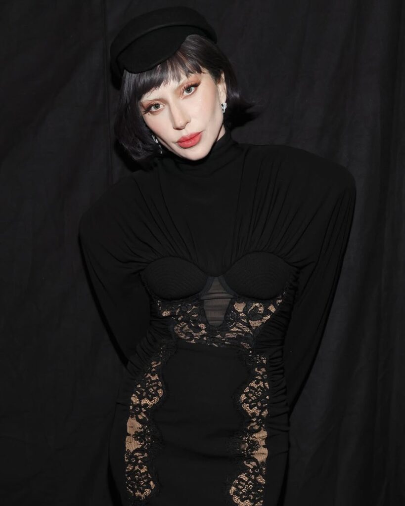 Lady Gaga with long black hair, large sunglasses, and a black catsuit flexes her bicep while standing behind four silver MTV VMA Moon Person trophies.