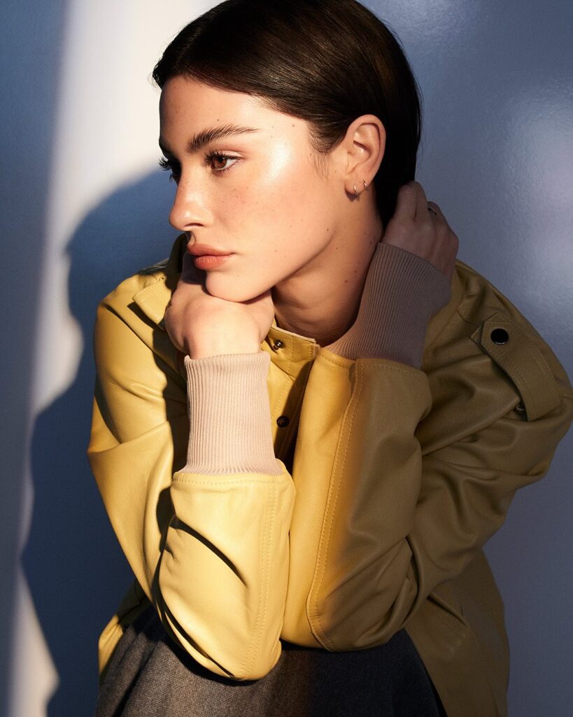 Profile portrait of  Gracie Abrams with dark hair in a bright yellow leather jacket and a ribbed beige cuff, resting her chin on her hands.