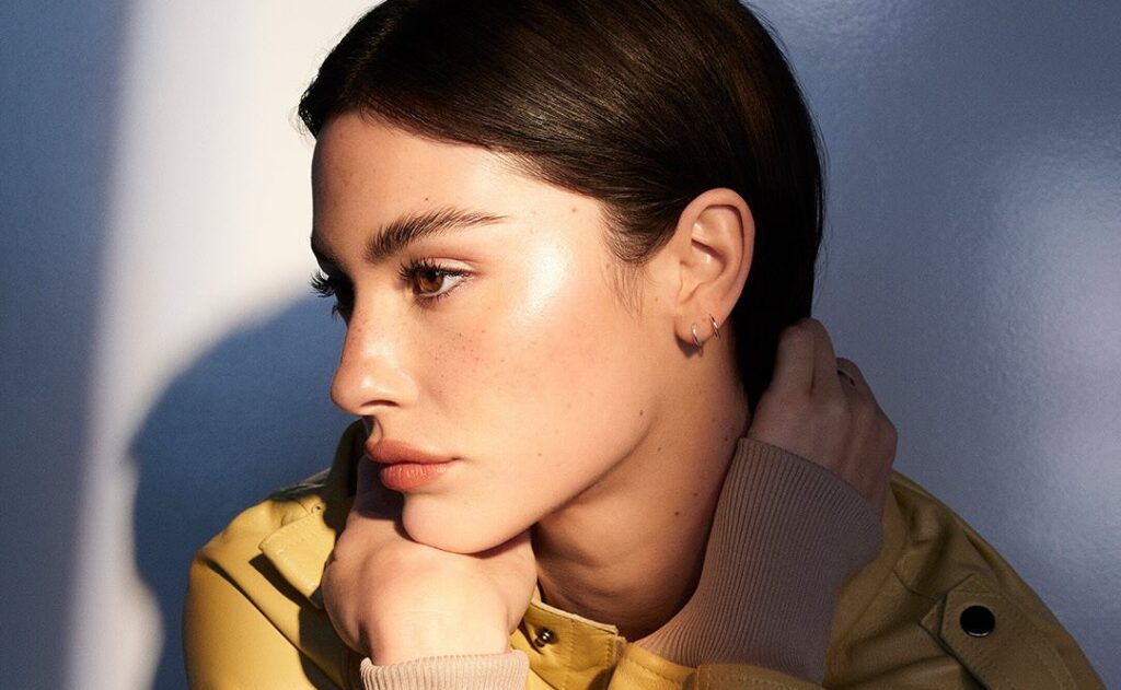 Profile portrait of Gracie Abrams with dark hair in a bright yellow leather jacket and a ribbed beige cuff, resting her chin on her hands.