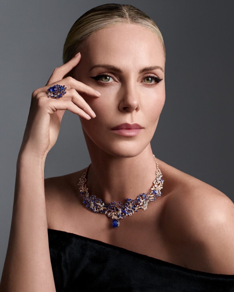 Elegant woman in an off‑shoulder black dress wearing a jeweled necklace and ring, resting her hand near her face against a gray studio background.