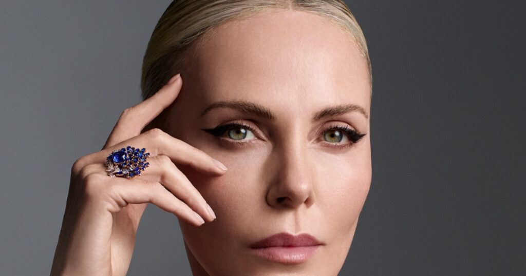 Elegant woman in an off‑shoulder black dress wearing a jeweled necklace and ring, resting her hand near her face against a gray studio background.