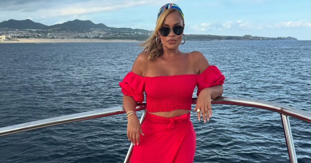 Gizelle Bryant standing on a yacht railing in a red off-shoulder top and skirt with ocean backdrop.