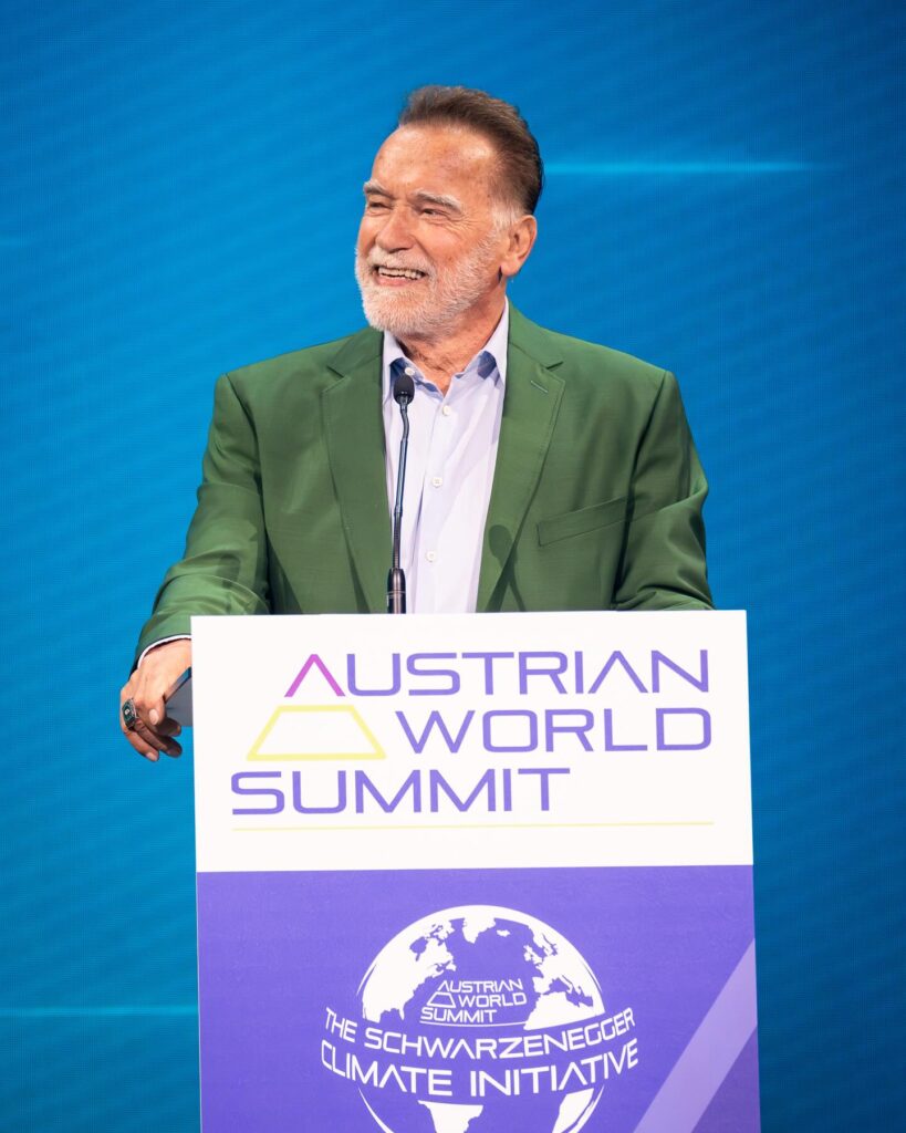 Full body shot of Arnold Schwarzenegger smiling with arms open, standing in front of a hedge and a large Netflix billboard that reads "FUBAR HAS B.D.E. (BIG DAD ENERGY)."