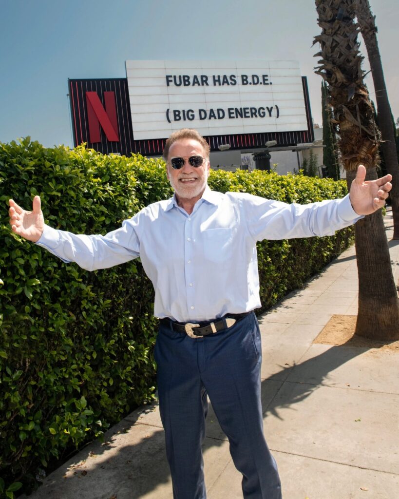 Arnold Schwarzenegger, wearing a green blazer, smiles while speaking at a podium during the Austrian World Summit. The podium logo is "AUSTRIAN WORLD SUMMIT."