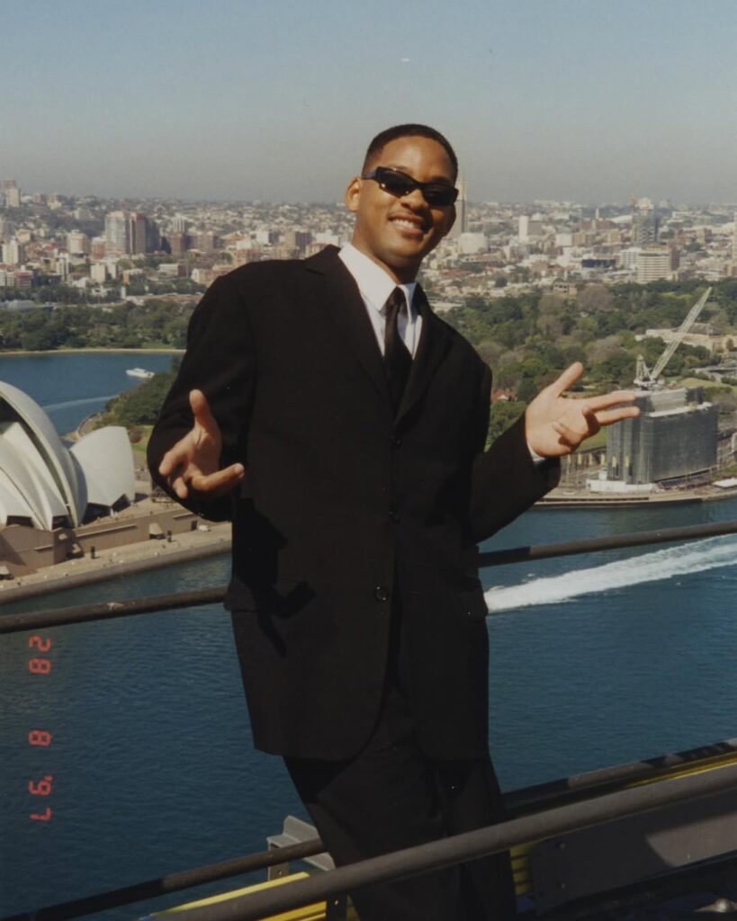 Will Smith performing live on stage at night, wearing a purple "West Philly" baseball jersey and cap, pointing into the crowd.