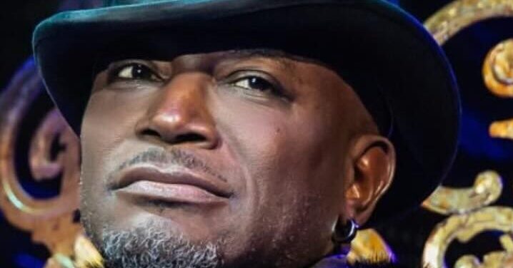 Taye Diggs in ornate dark brocade coat with fur collar and tall top hat, standing before decorative gilded set pieces, looking confidently off-camera.