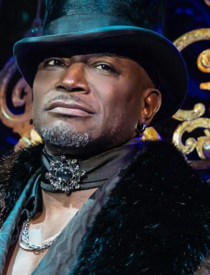 Taye Diggs in ornate dark brocade coat with fur collar and tall top hat, standing before decorative gilded set pieces, looking confidently off-camera.