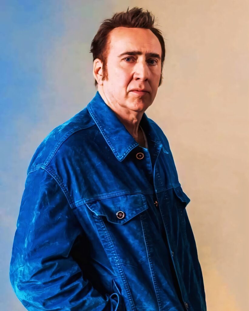 Nicolas Cage a blue jacket standing against a neutral, two‑tone background, looking toward the camera.