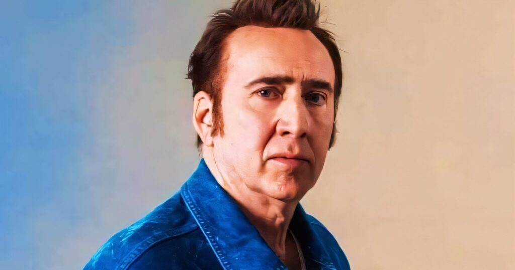 Nicolas Cage wearing a blue jacket standing against a neutral, two‑tone background, looking toward the camera.