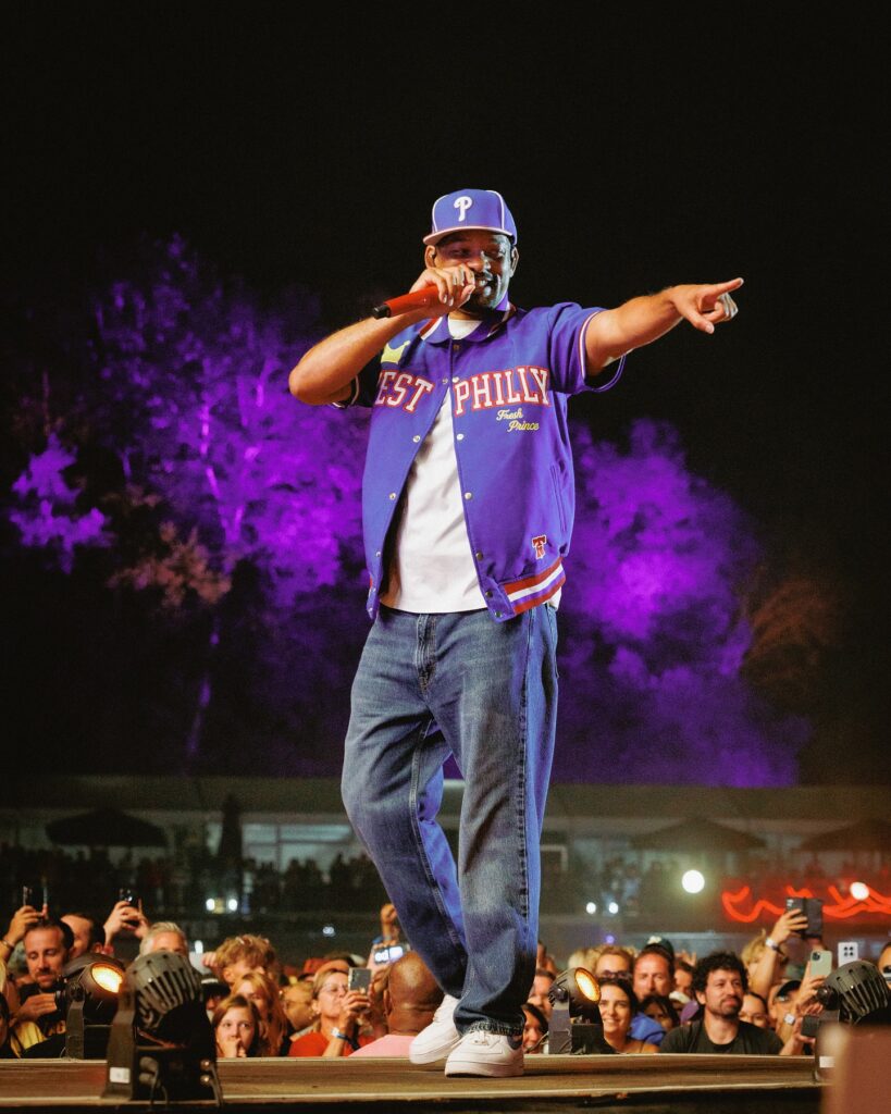 Will Smith smiling in a black suit and sunglasses, posing on a railing with the Sydney skyline and Opera House in the background.