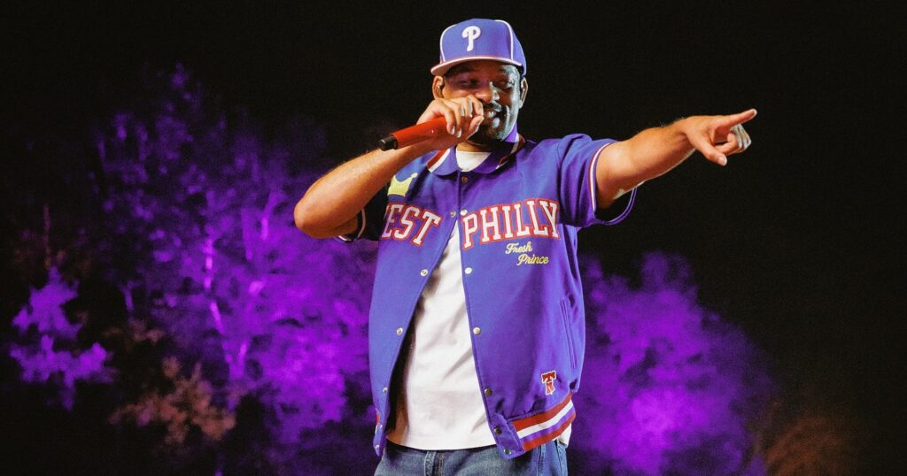 Will Smith smiling in a black suit and sunglasses, posing on a railing with the Sydney skyline and Opera House in the background.