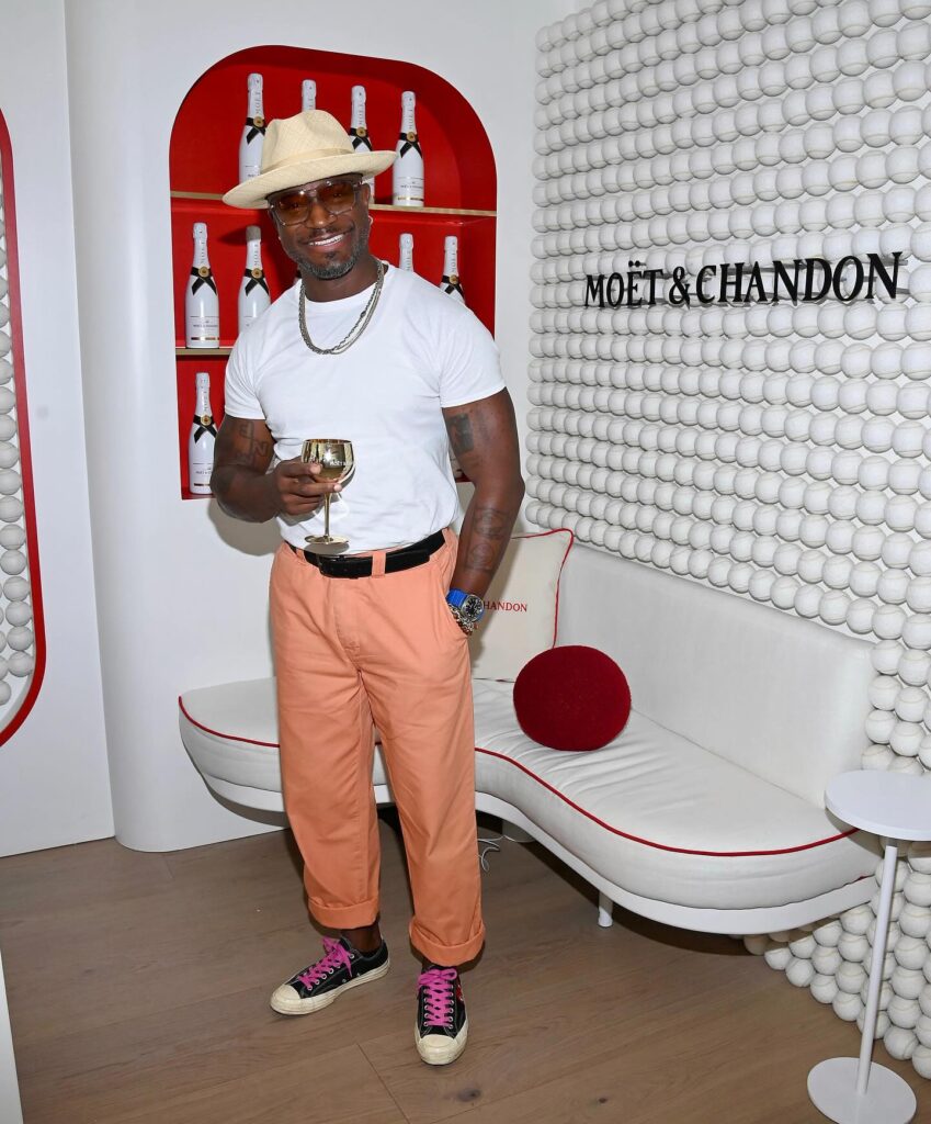 Taye Diggs in white tee, peach trousers, straw hat and pink-laced sneakers holding a drink while posing in a branded lounge with bottle display.