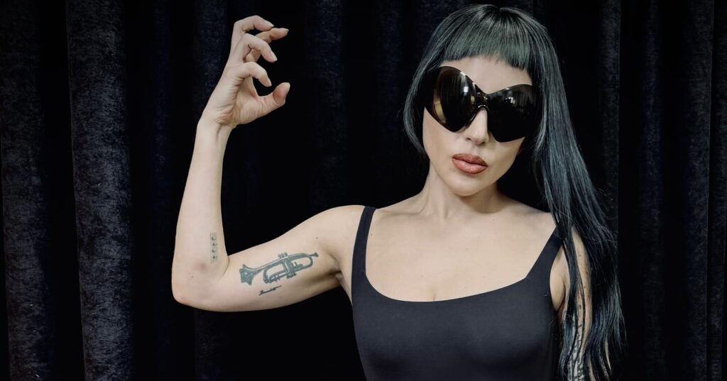 Lady gaga in a short black bob and newsboy cap, wearing a black high-neck dress featuring a structured bustier and lace paneling.
