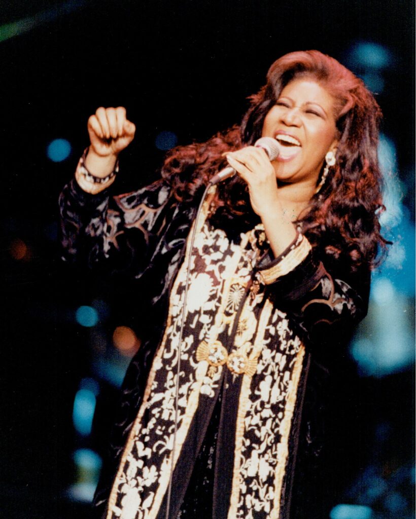 Live performance photo of a woman singing passionately into a microphone, wearing a patterned long coat.
